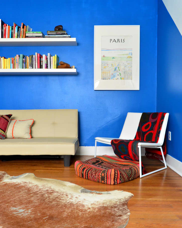 Blue-walled room with a beige sofa, colorful bookshelves, Paris poster, patterned chair, and cowhide rug on wooden floor.