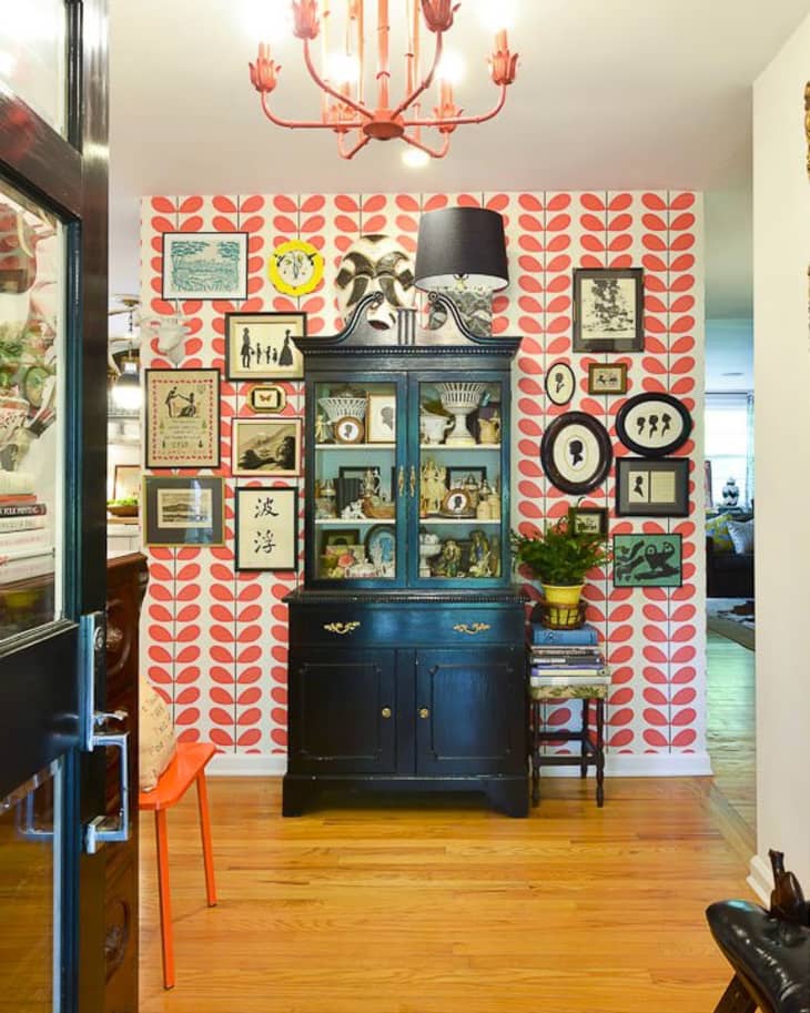 Eclectic hallway with red patterned wallpaper, black cabinet, framed art, red chandelier, and orange chair.
