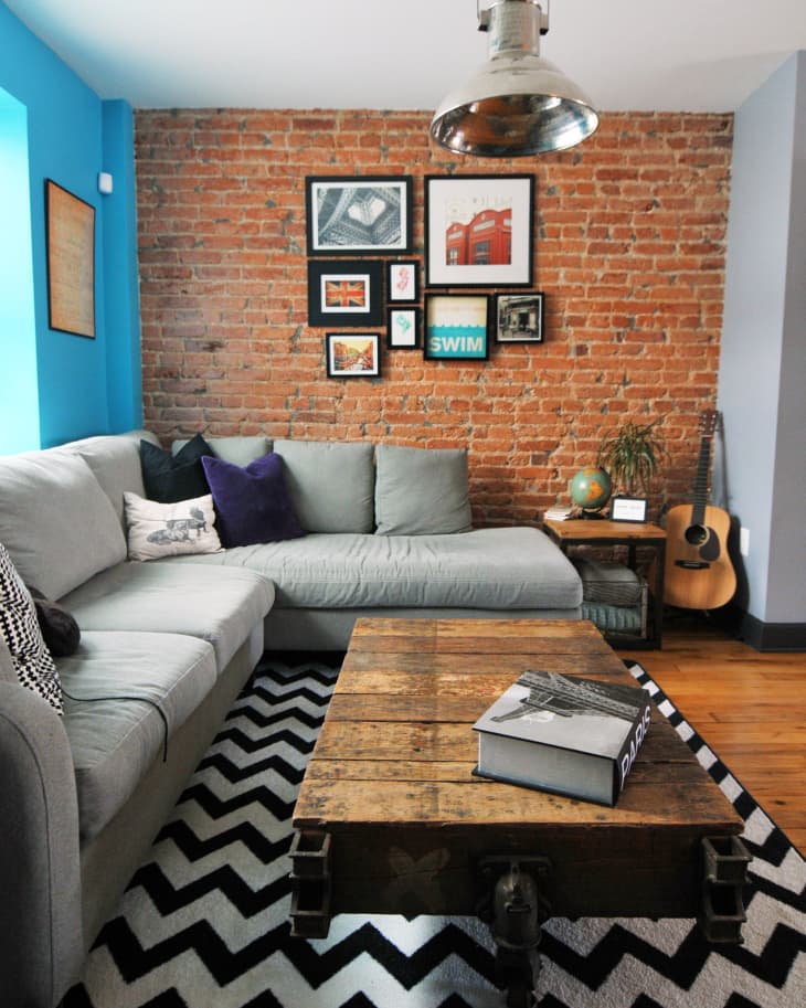 Living room with gray sectional sofa, brick wall, framed art, rustic coffee table, chevron rug, and a guitar in the corner.