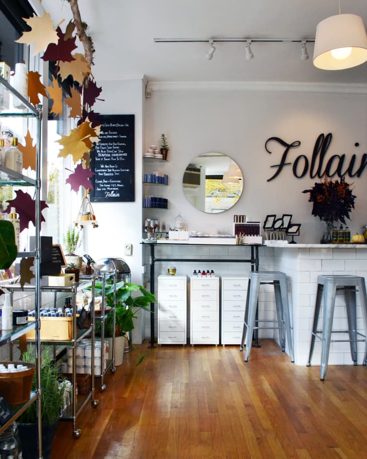 Store interior with wooden floors, metal shelves, a round mirror, and stools at a white counter. Autumn leaves hang from a branch.