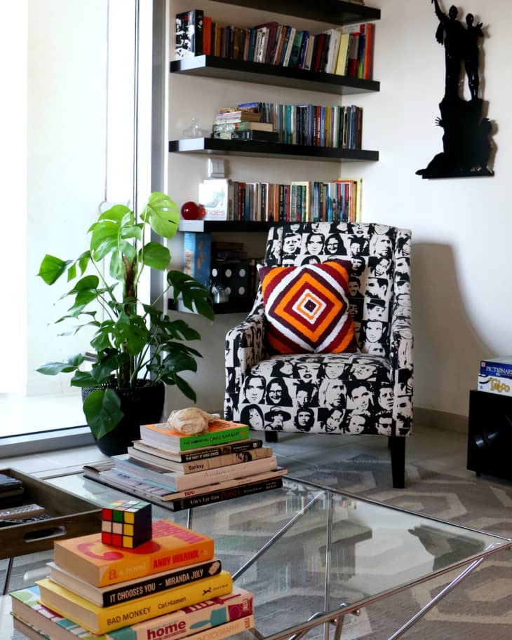 Living room with patterned armchair, colorful cushion, glass coffee table, books, Rubik's cube, and potted plant.