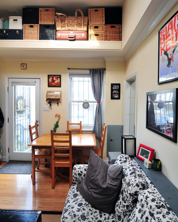 Living room with floral sofa, wooden dining table, chairs, and wall art. Storage baskets and suitcase on upper shelf.