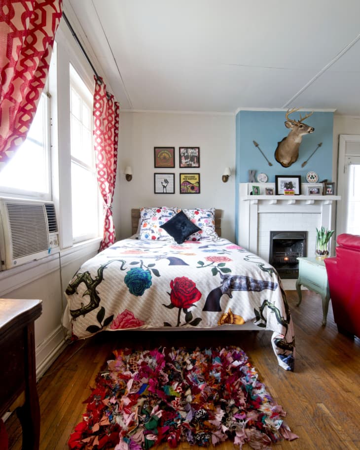 Bedroom with floral bedspread, colorful rug, red curtains, and a deer head mounted above a white fireplace.