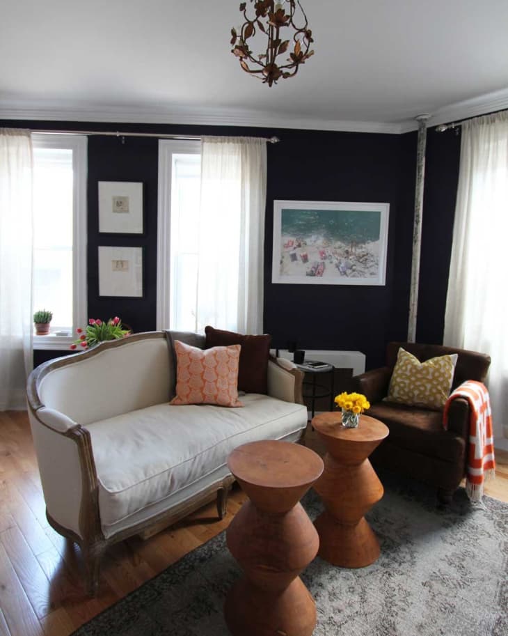 Living room with a cream sofa, brown armchair, wooden tables, floral chandelier, and dark walls.