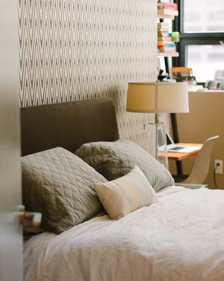 Cozy bedroom with patterned wallpaper, a bed with green pillows, a lamp, and a small desk by the window.