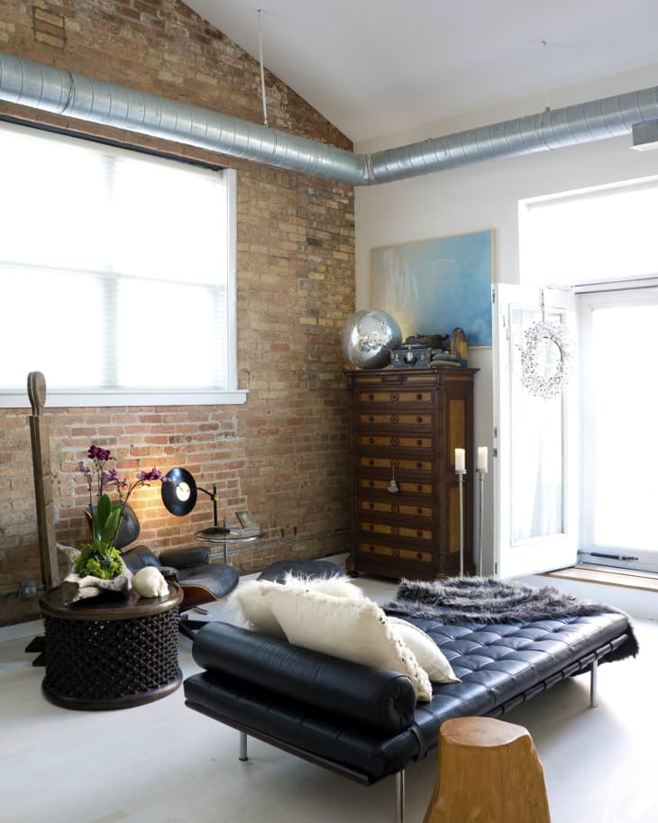 Loft-style living room with exposed brick, black leather daybed, wooden stool, and vintage chest of drawers.