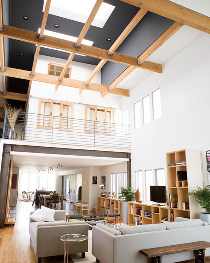 Spacious living room with high ceiling, skylights, gray sofas, wooden shelves, and dining area in the background.