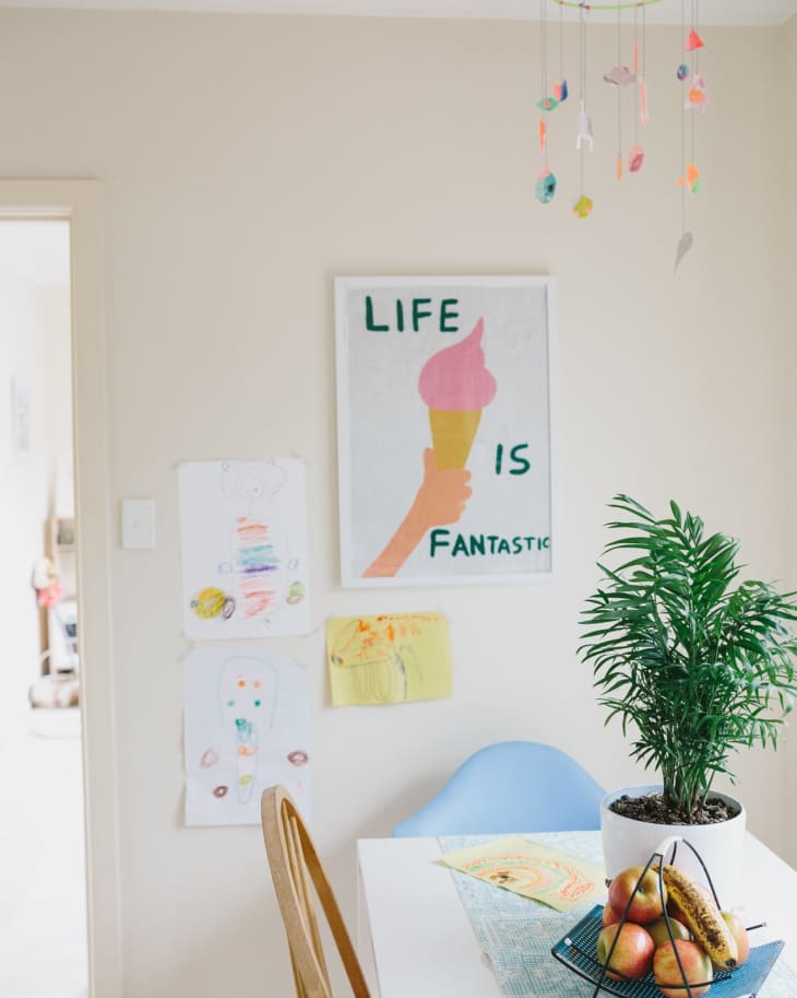 Dining area with a white table, fruit basket, potted plant, colorful mobile, and wall art saying "Life is Fantastic."
