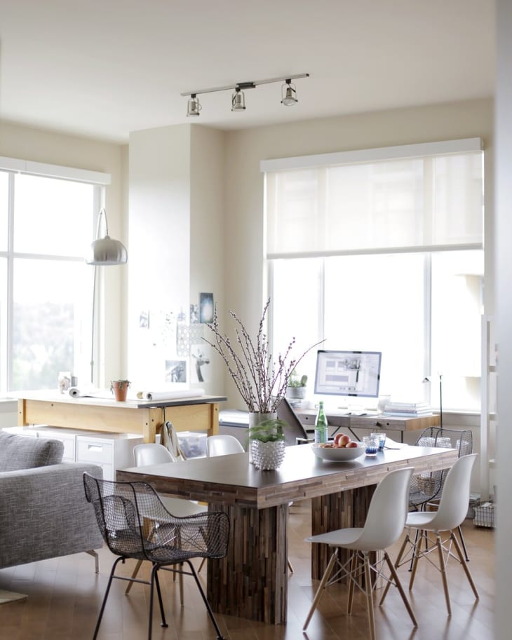 Modern dining area with wooden table, mixed chairs, vase with branches, and a computer on a desk by large windows.