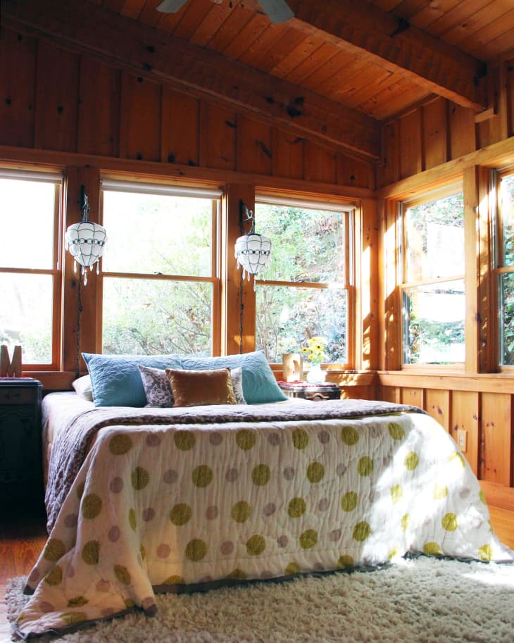 Cozy bedroom with wood paneling, large windows, polka dot bedspread, and hanging pendant lights.