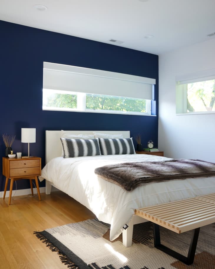 Modern bedroom with navy accent wall, white bed, striped pillows, wooden nightstand, and bench on a patterned rug.
