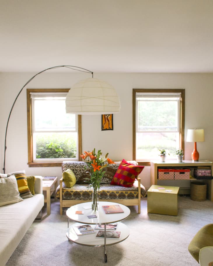 Living room with floral sofa, colorful cushions, round coffee table, and large floor lamp. Orange lilies in a vase.