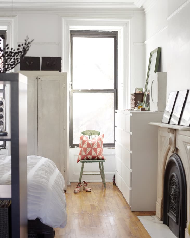 Bedroom with a green chair, red geometric pillow, white dresser, and fireplace, featuring hardwood floors and a large window.