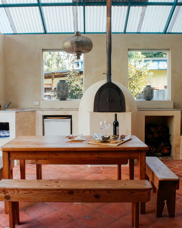Wooden dining table with benches, set with wine and snacks, in a rustic kitchen with a pizza oven and pendant light.