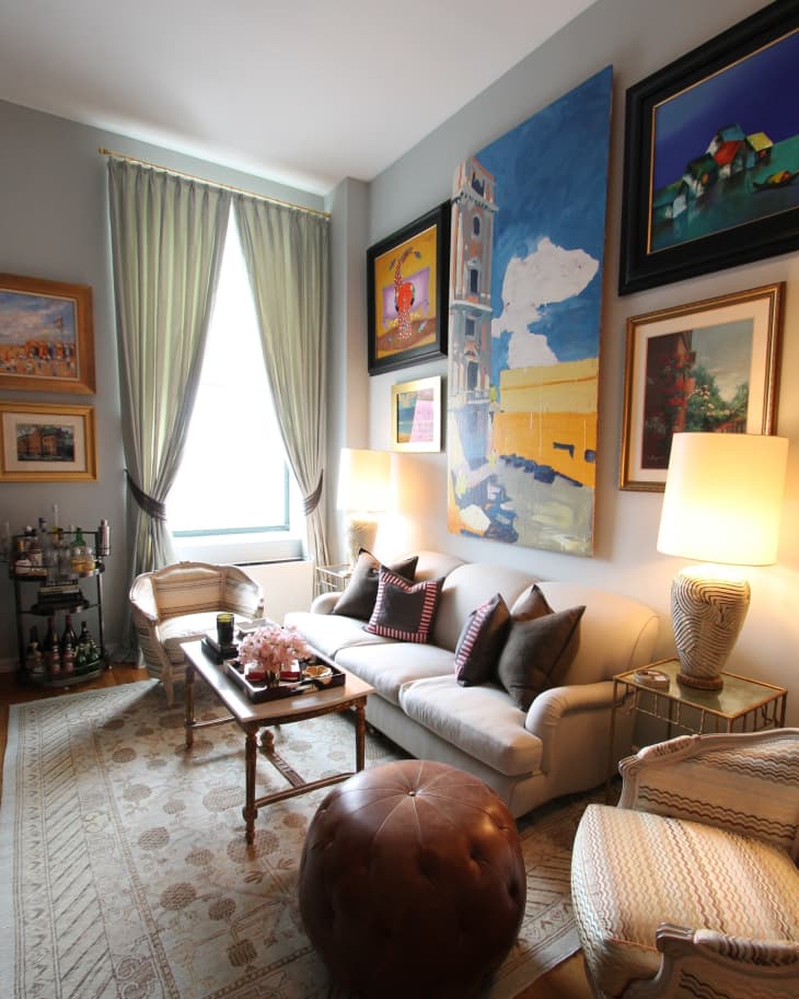 Living room with a white sofa, colorful pillows, large paintings, a bar cart, and a round leather ottoman.
