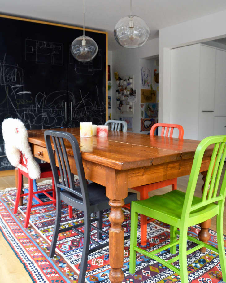Dining room with wooden table, colorful chairs, patterned rug, and chalkboard wall.