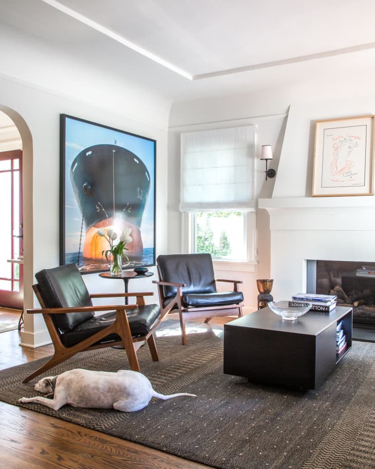 Living room with black leather chairs, a large ship painting, a dog on a rug, and a modern coffee table with books.