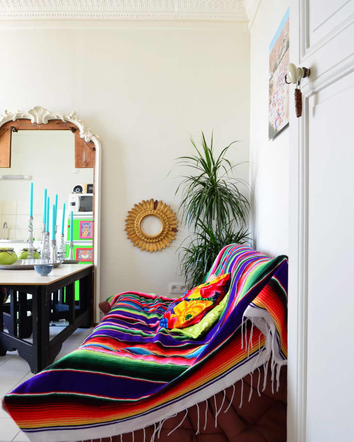 Colorful striped blanket on a sofa, with a sunflower pillow, next to a large plant and a decorative mirror.