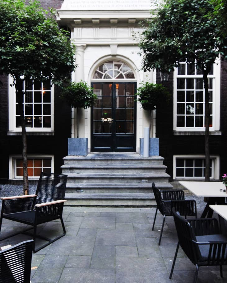Courtyard with black chairs, stone steps, and arched doorway flanked by large windows and potted trees.