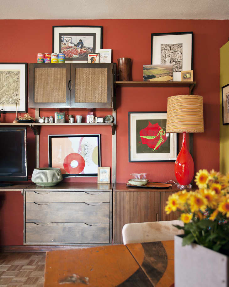 Mid-century modern living room with red walls, wooden cabinets, framed art, a red lamp, and a table with yellow flowers.