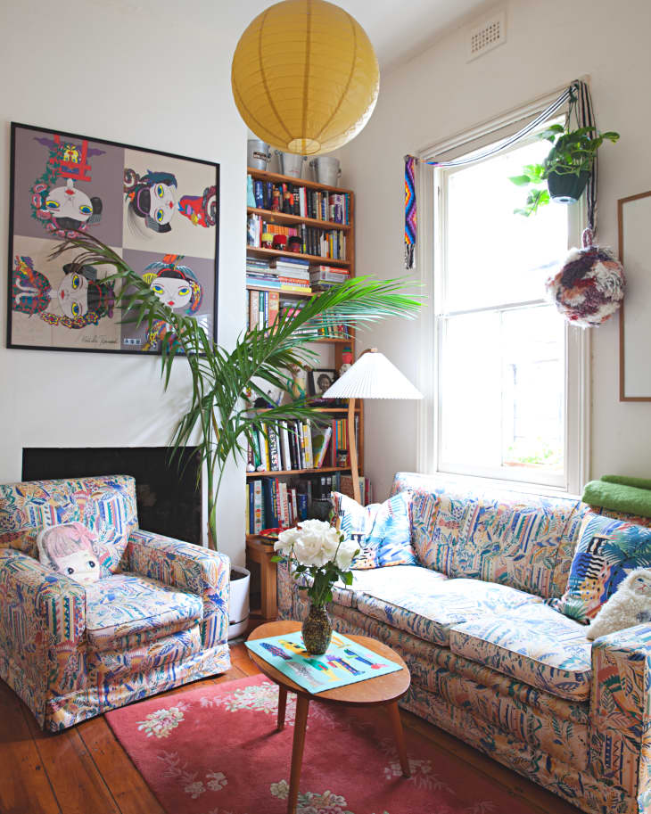Cozy living room with patterned sofa and armchair, red rug, bookshelf, potted plant, and large yellow paper lantern.