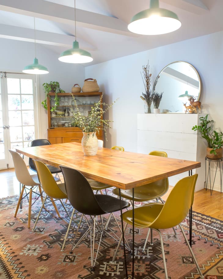 Dining room with wooden table, mixed chairs, patterned rug, pendant lights, and plants on a sideboard.