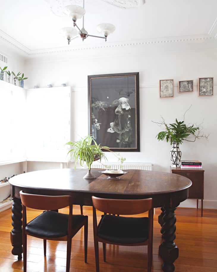 Dining room with dark wood table, two chairs, potted plants, and framed artwork on the wall.