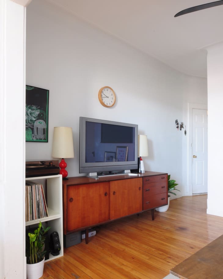 Mid-century modern living room with wooden sideboard, TV, red lamps, vinyl records, and potted plants on hardwood floor.