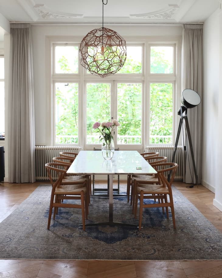 Dining room with a marble table, six wooden chairs, a geometric pendant light, and a vase of flowers on a patterned rug.