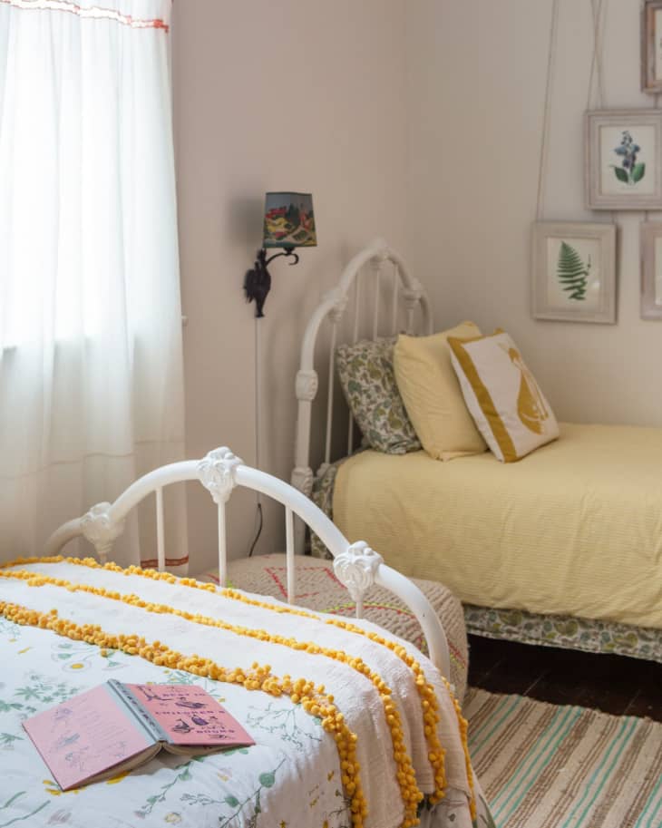 Cozy bedroom with two white metal beds, yellow bedding, floral prints, and a pink book on the bed.