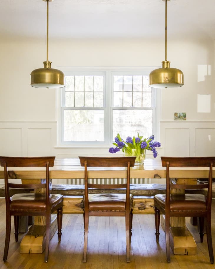 Dining room with wooden table, four chairs, brass pendant lights, and a vase of purple flowers by the window.