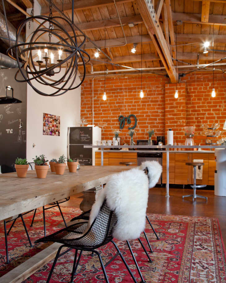Loft kitchen with exposed brick, wooden beams, industrial chandelier, and a large wooden table with potted plants.