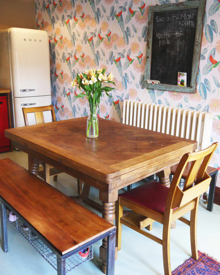 Dining area with wooden table, chairs, bench, floral wallpaper, chalkboard, and vase of flowers. White fridge in the corner.