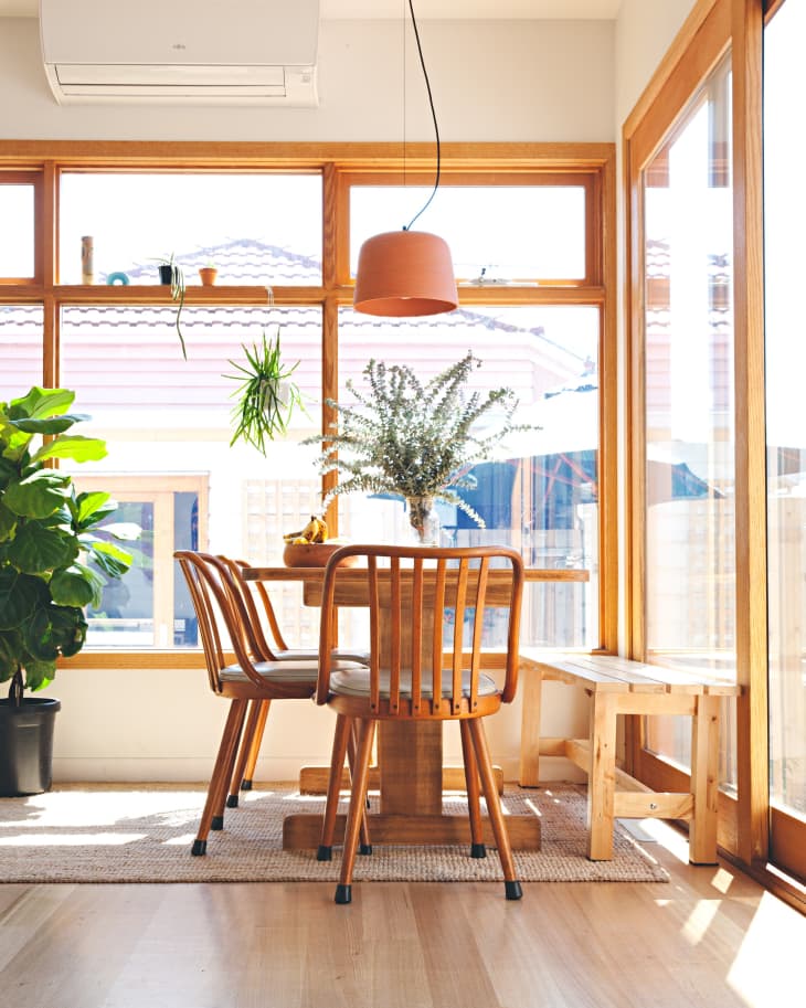 Sunlit dining area with wooden table, chairs, bench, hanging plants, and large windows.