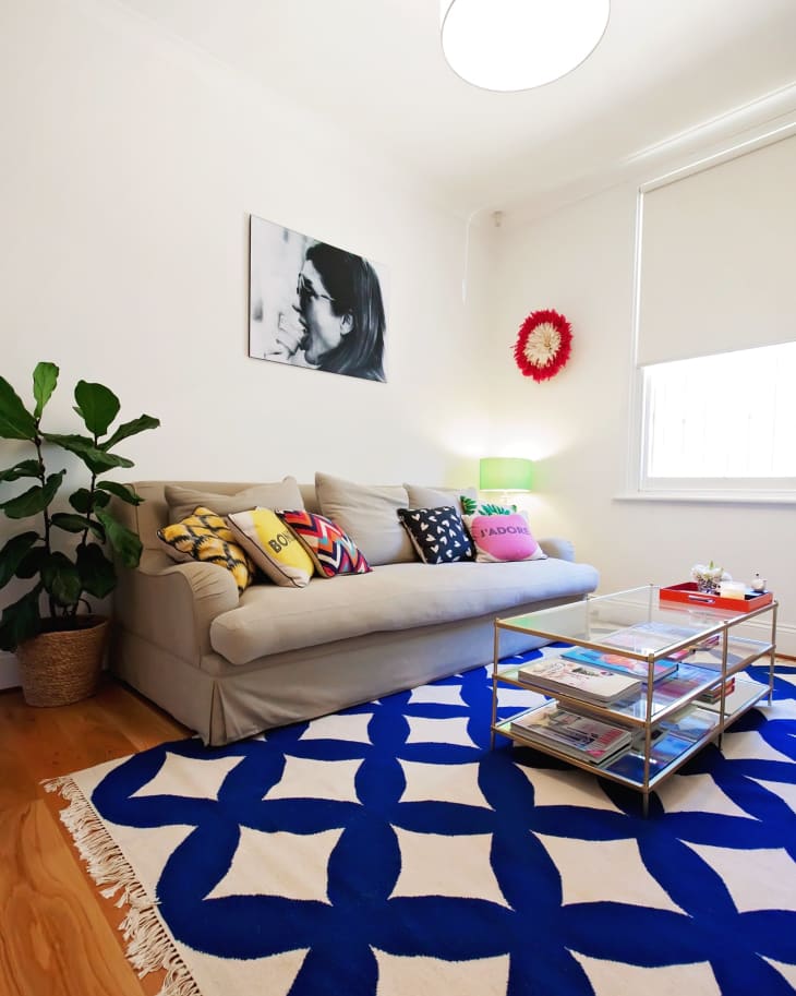 Living room with beige sofa, colorful pillows, blue patterned rug, glass coffee table, and potted plant.