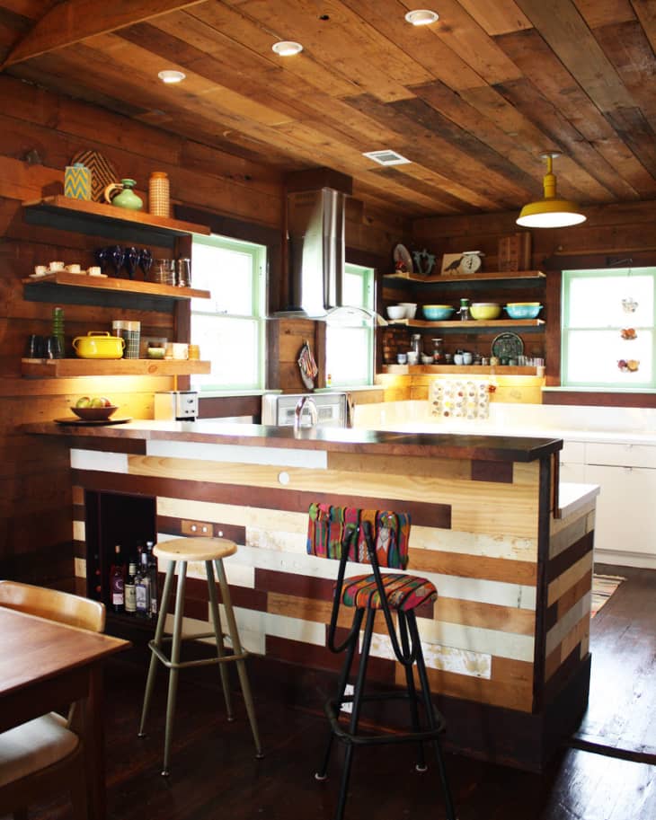 Rustic kitchen with wooden walls, open shelves, colorful dishes, striped bar stools, and a wooden ceiling with recessed lighting.