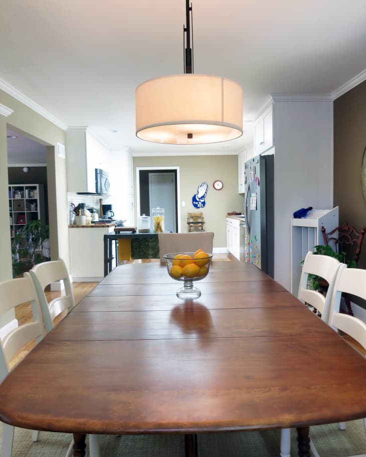 Dining room with a wooden table, white chairs, pendant light, and a bowl of oranges, leading to a kitchen area.