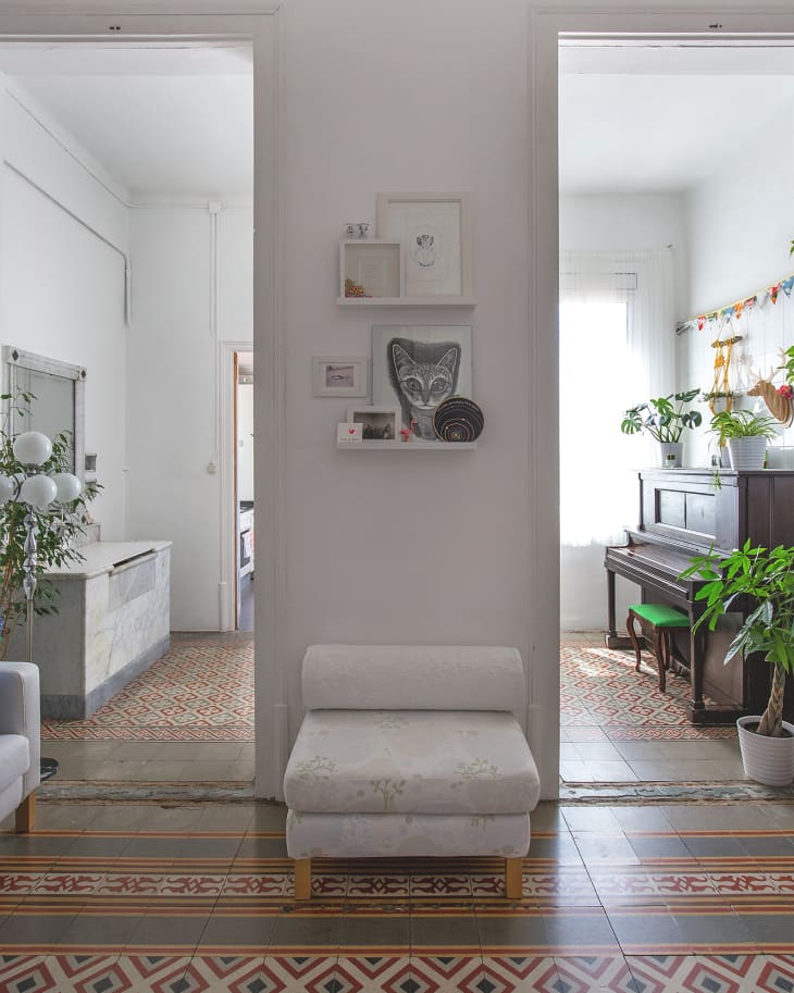 White chair in hallway with patterned floor tiles, wall shelves, and view into rooms with plants and a piano.