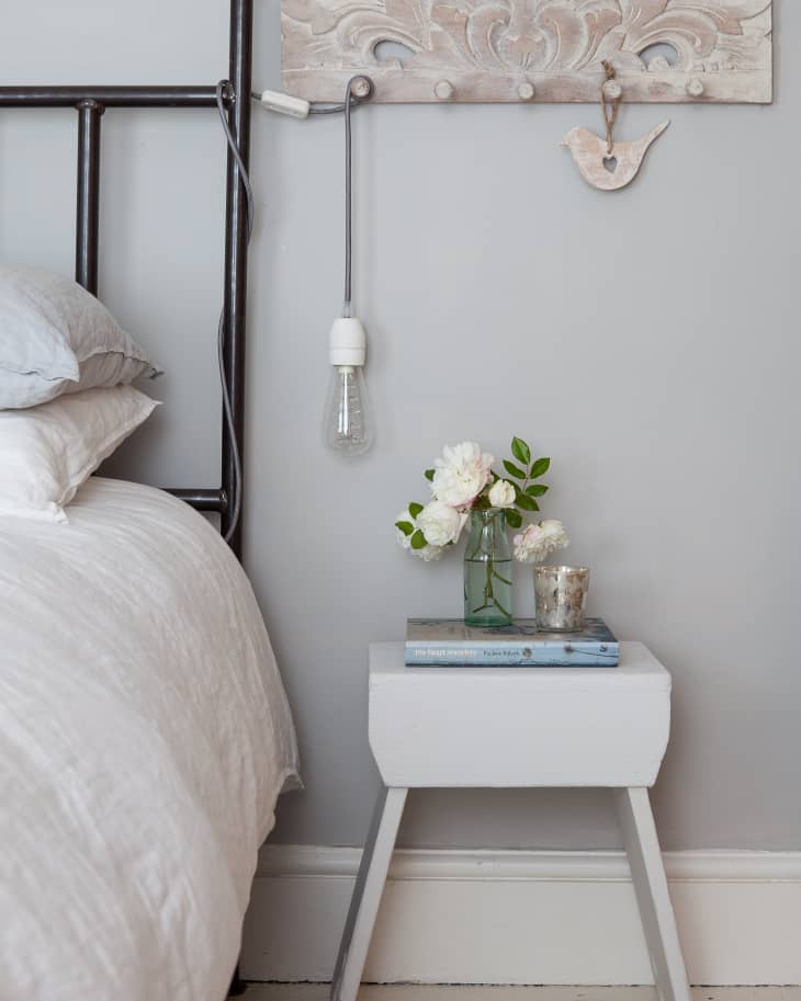 Bedside table with flowers in a jar, a book, and a candle, next to a bed with a metal frame and a hanging light bulb.