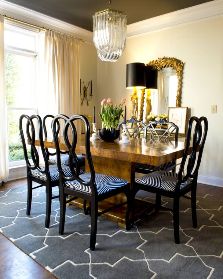 Dining room with wooden table, black chairs, striped cushions, chandelier, and gold mirror on a gray patterned rug.