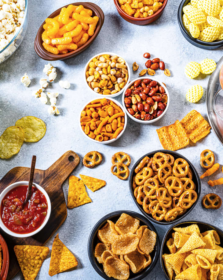 Party food: assortment of salty snacks in bowls and drinks like beer, cola and cocktail shot from above on party table. The composition includes potato chips, popcorn, nachos and salsa, corn bugles, pretzels, peanut, pistachio, cheese sticks and drinks like beer, cola soda and cocktail. Predominant colors are yellow and gray. High resolution 42Mp studio digital capture taken with SONY A7rII and Zeiss Batis 40mm F2.0 CF lens