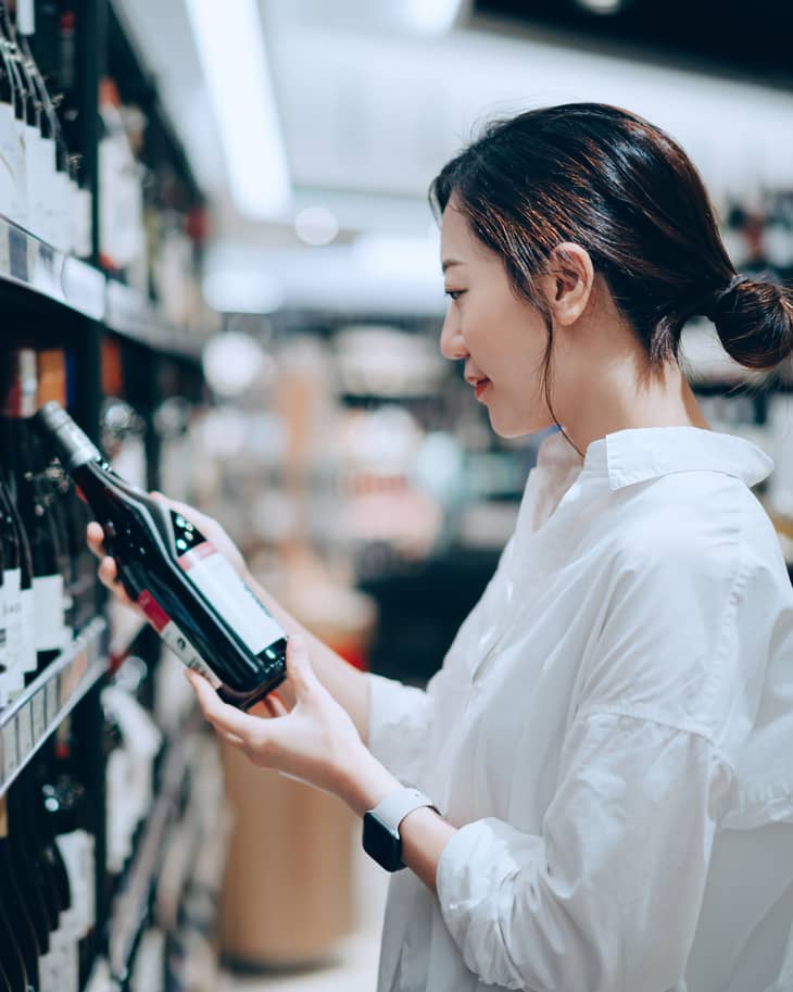 Young Asian woman walking through liquor aisle and choosing a bottle of red wine from the shelf in supermarket