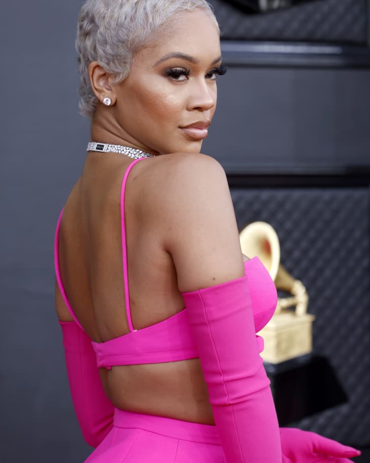 Rapper Saweetie at the 2022 Grammy Awards with short blonde hair, wearing neon pink top, gloves. Looking back at the camera over her shoulder