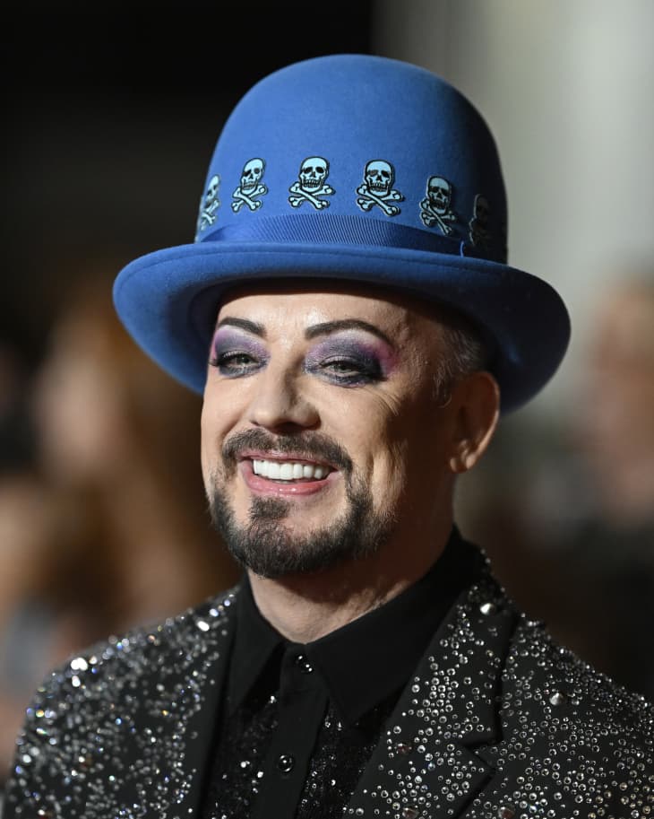 Photo portrait of Boy George wearing a blue hat and sequined jacket, smiling at an event