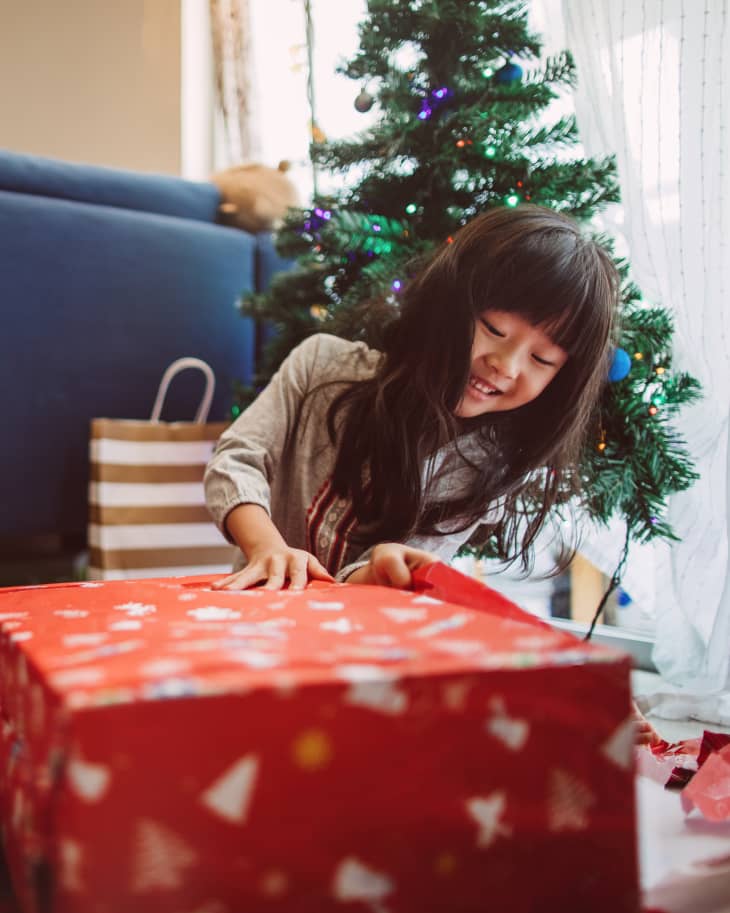 Child opening a gift in front of a christmas tree. Other gifts and blue sofa in background