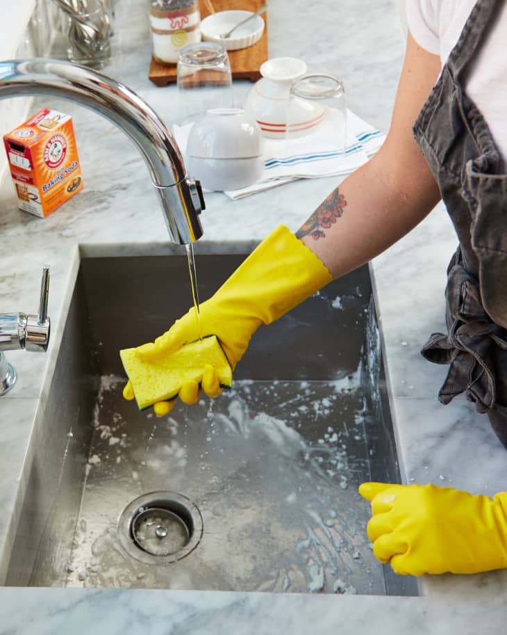 someone cleaning a stainless steel kitchen sink with a sponge and baking soda. Dishes are drying on towels on the counter