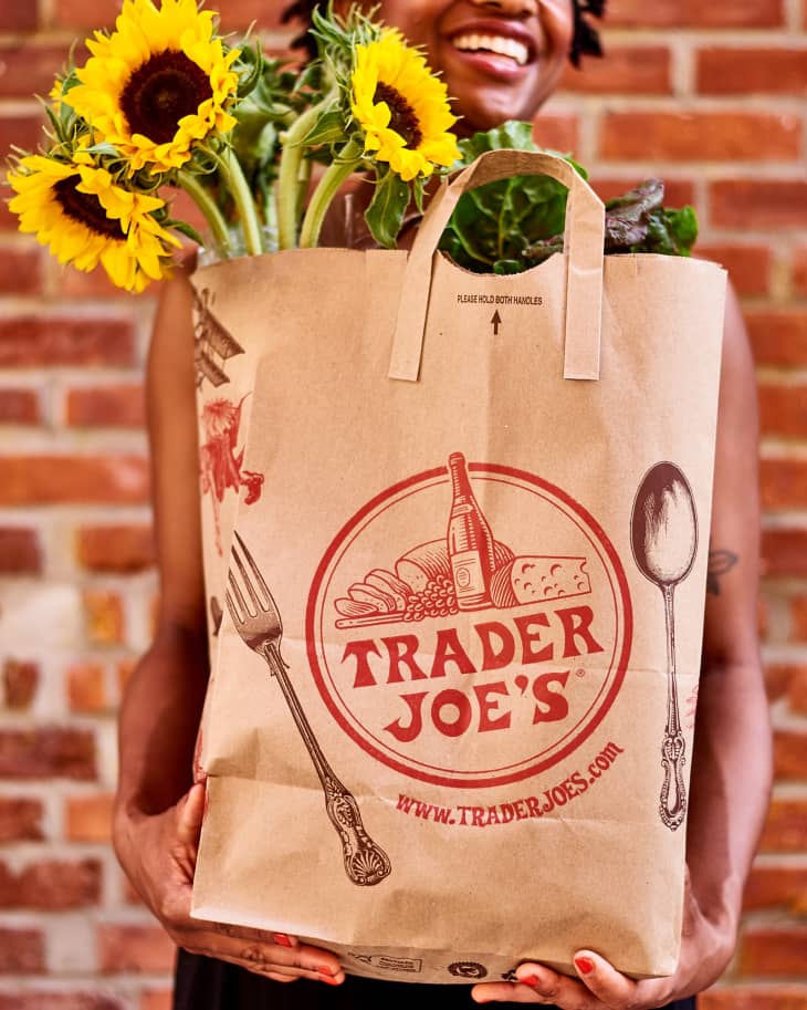 Woman holding trader joe's paper bag with both hands and smiling. Sunflowers sticking out the top