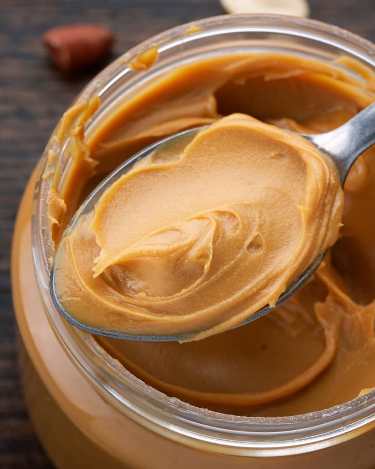 peanut butter in an open jar with spoon and peanuts scattered on the table