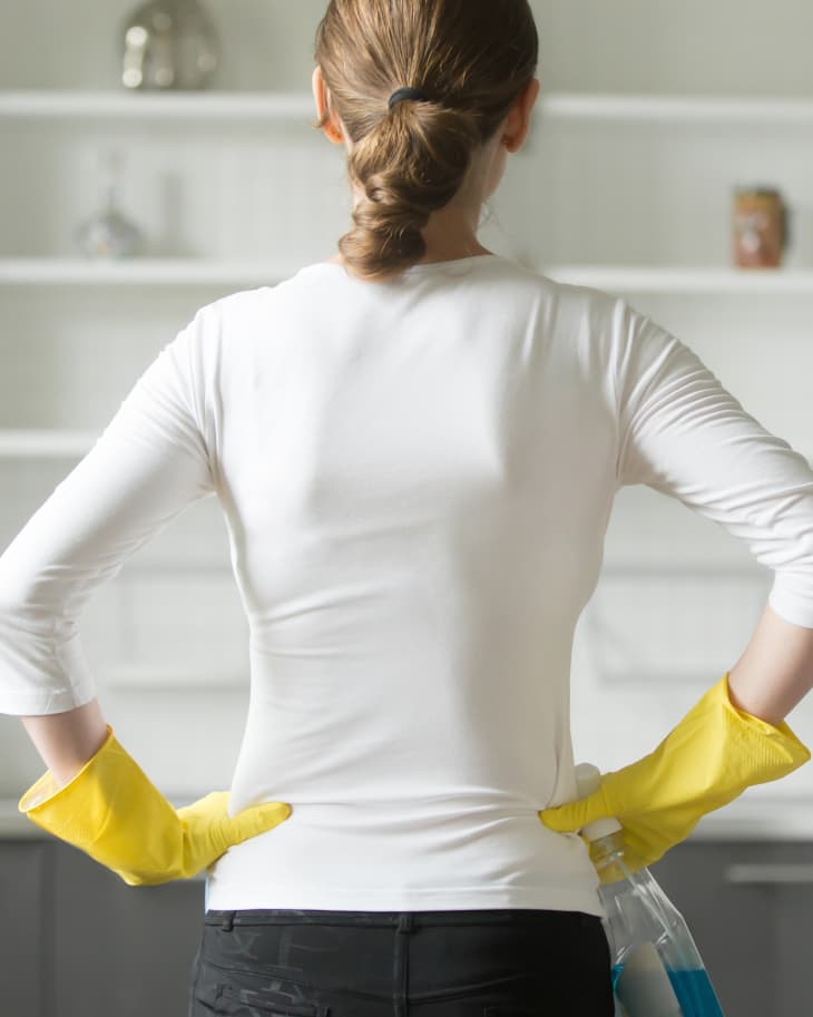 Rear view of young woman wearing rubber protective yellow gloves, holding rag and spray bottle detergent, hands at her hips, observing clean and sparkling kitchen.