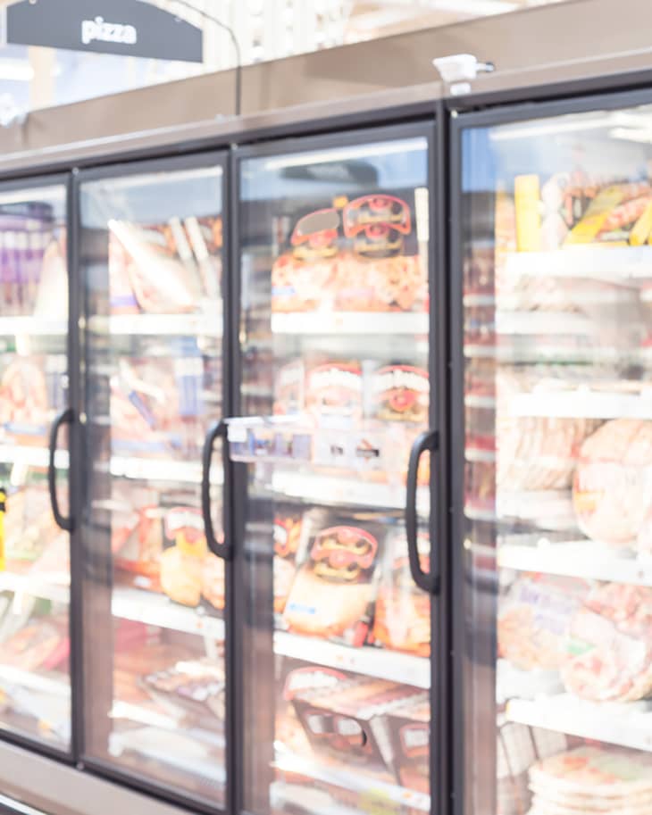 Blurred frozen food section at retail store in America. Huge glass door aisle with variety pack of processed pizza. Freezer full assortment of frozen pizza in local supermarket, defocused background.